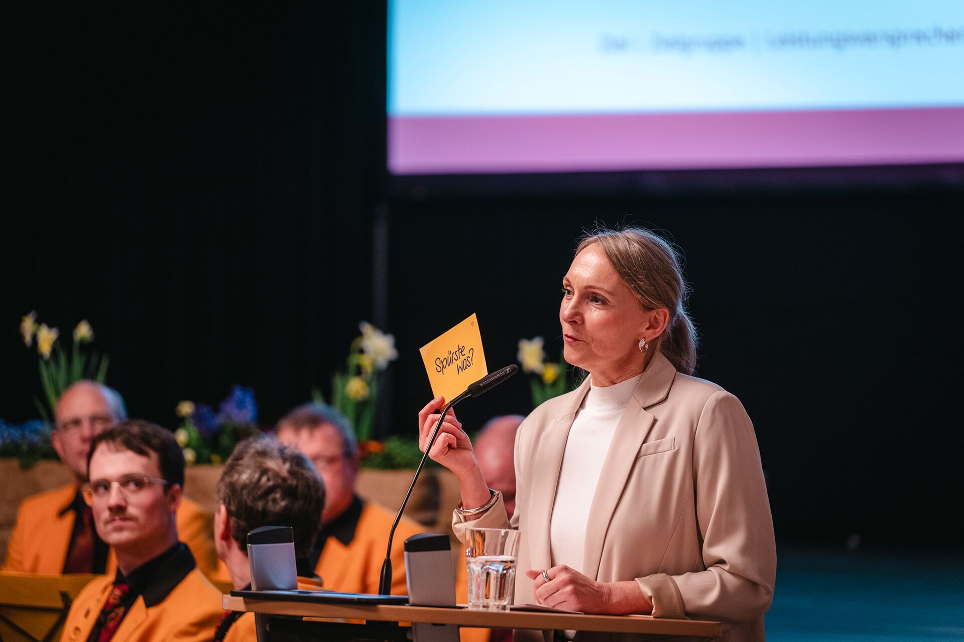 Maren Thier am Rednerpult zeigt gelbe Karte mit blauem Schriftzug „Spüste was?”