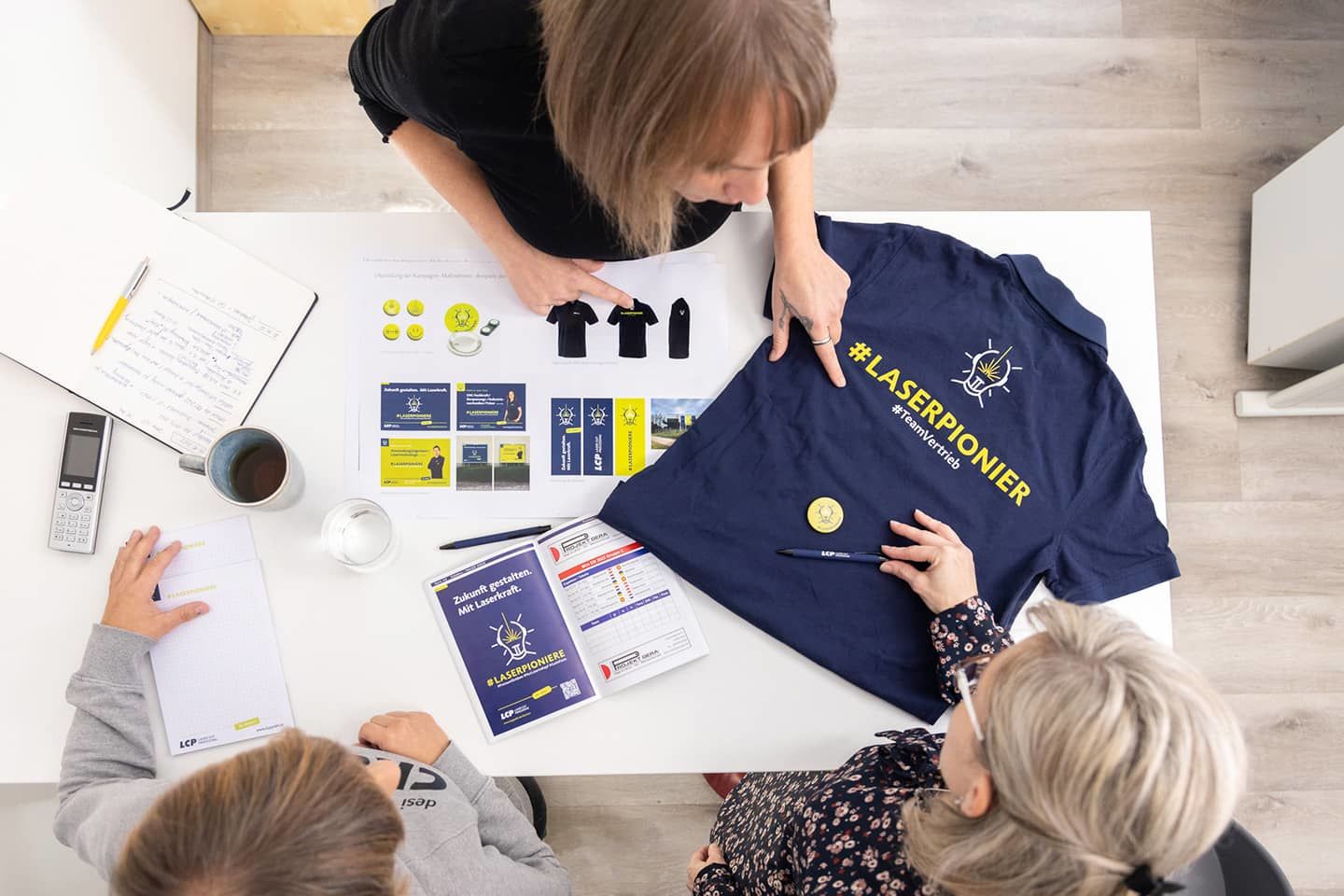 Blick von oben auf das Recruiting-Kampagnen-Design: Anzeigen, Moodboard und Mitarbeiter-T-Shirts mit gelbem Schriftzug #Laserpionier