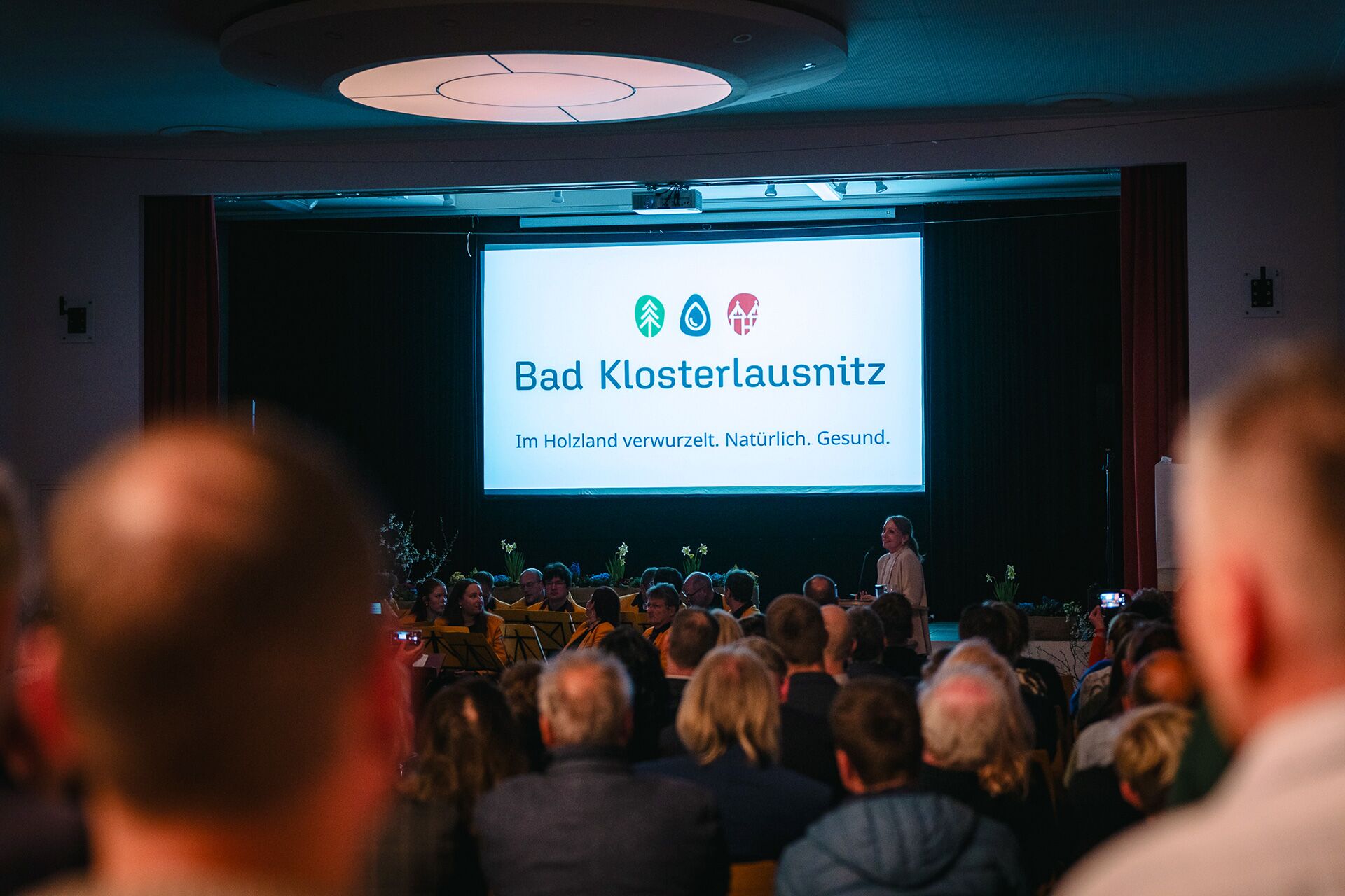 Maren Thier am Rednerpult präsentiert die neuen Markenstrategie vor Leinwand mit dem neuen Logo von Bad Klosterlausnitz, Zuschauerblick über die Schulter
