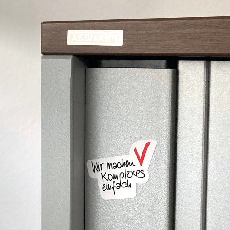 Zukunftsbild-Sticker „Wir machen komplexes einfach” mit rotem Häkchen, geklebt auf Ordnerrücken